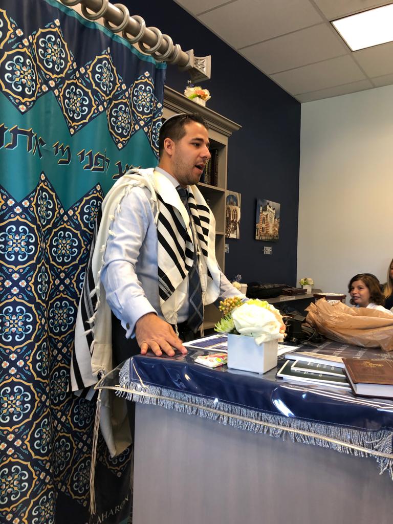 A teacher in a tallit leading a lesson beside the Torah ark at the chapter's Beit Midrash.